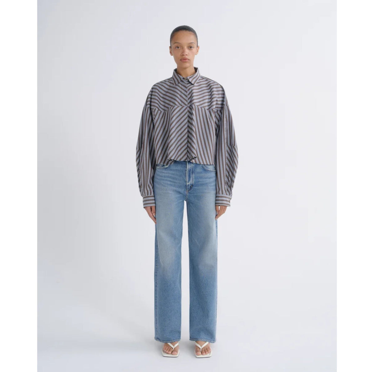 Full-length front view of a model wearing a striped long-sleeve collared shirt tucked into high-waisted AGOLDE Ren Jeans in Measure with light brown heels against a light gray background.