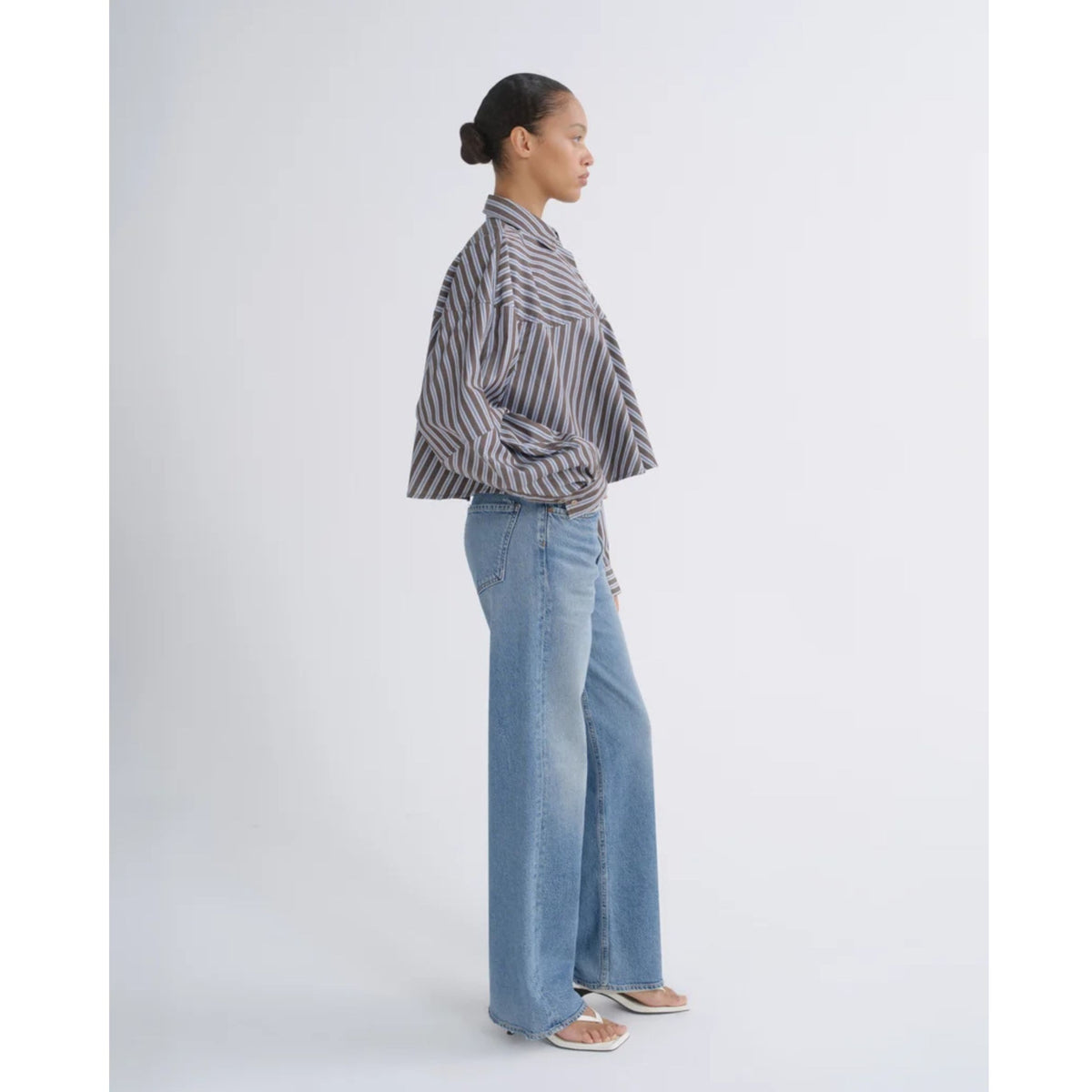 Side profile view of a model wearing a striped button-down shirt and AGOLDE Ren Jeans in Measure against a light gray background.