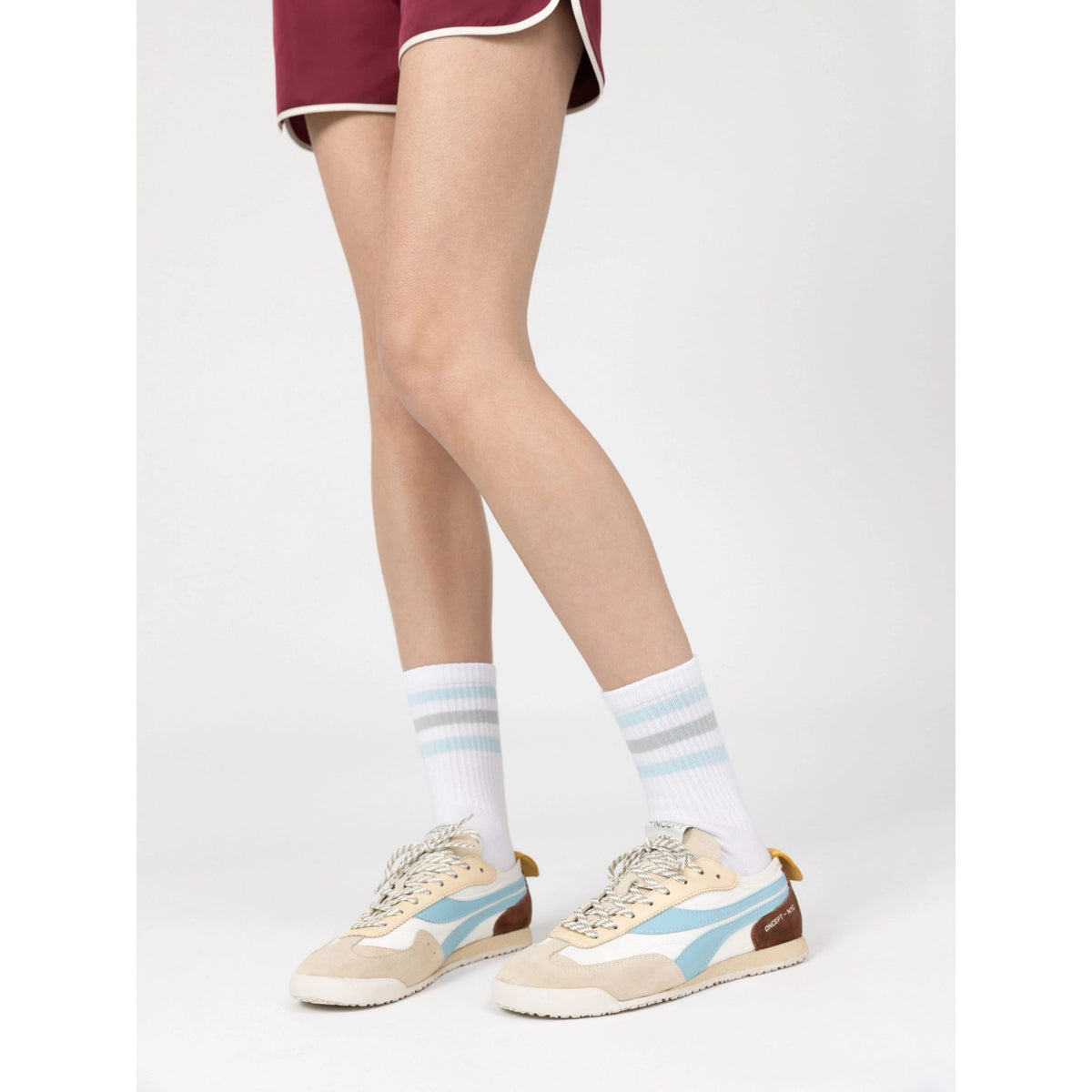 A person from mid-thigh down wearing white crew socks with light blue and gray stripes, paired with beige and light blue sneakers, against a plain light gray studio background.