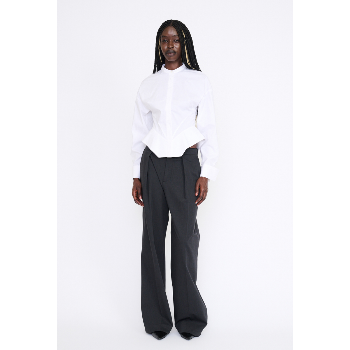 A model stands against a plain white studio background wearing a white long-sleeve cropped blouse with structured shoulders and dark charcoal wide-leg trousers.