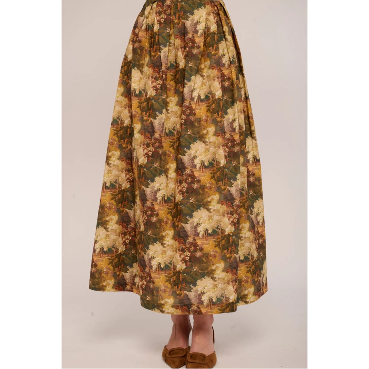 A high-waisted, voluminous A-line skirt with a vintage floral and landscape print in earthy tones of olive green, mustard yellow, and beige, shown on a model wearing brown flat shoes against a light beige studio background.