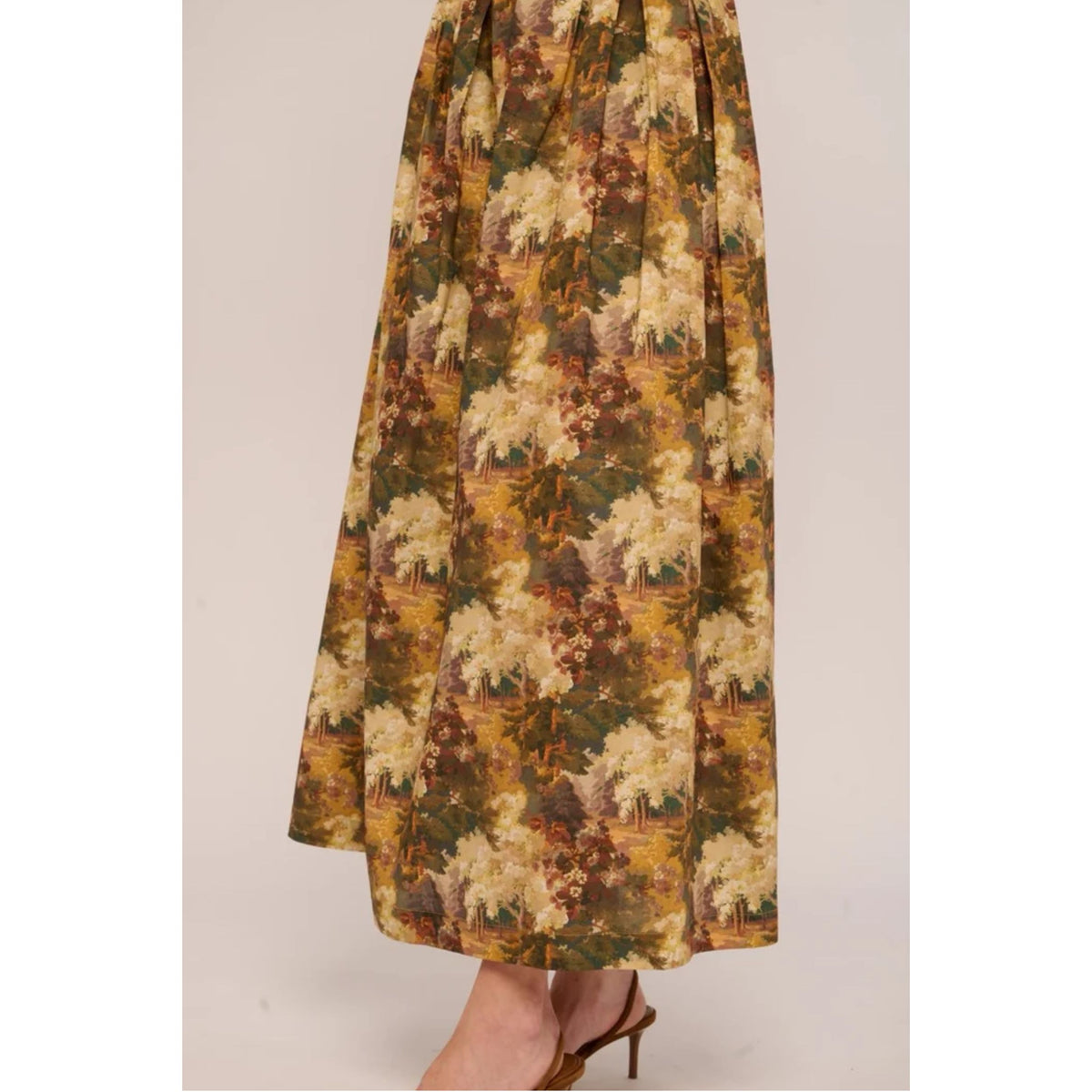 A high-waisted, floor-length skirt with a vintage floral landscape print in earthy olive green, mustard yellow, and cream tones, shown on a model wearing brown high-heeled sandals.