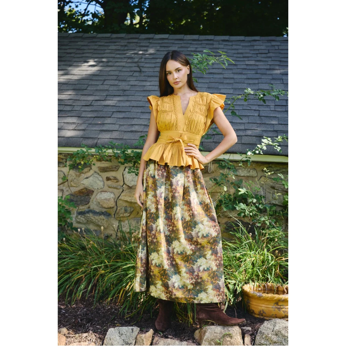 A woman stands outdoors in front of a stone wall and greenery, wearing a mustard yellow ruffled blouse with short flutter sleeves and a matching long floral skirt in yellow, beige, and brown tones, paired with brown boots.