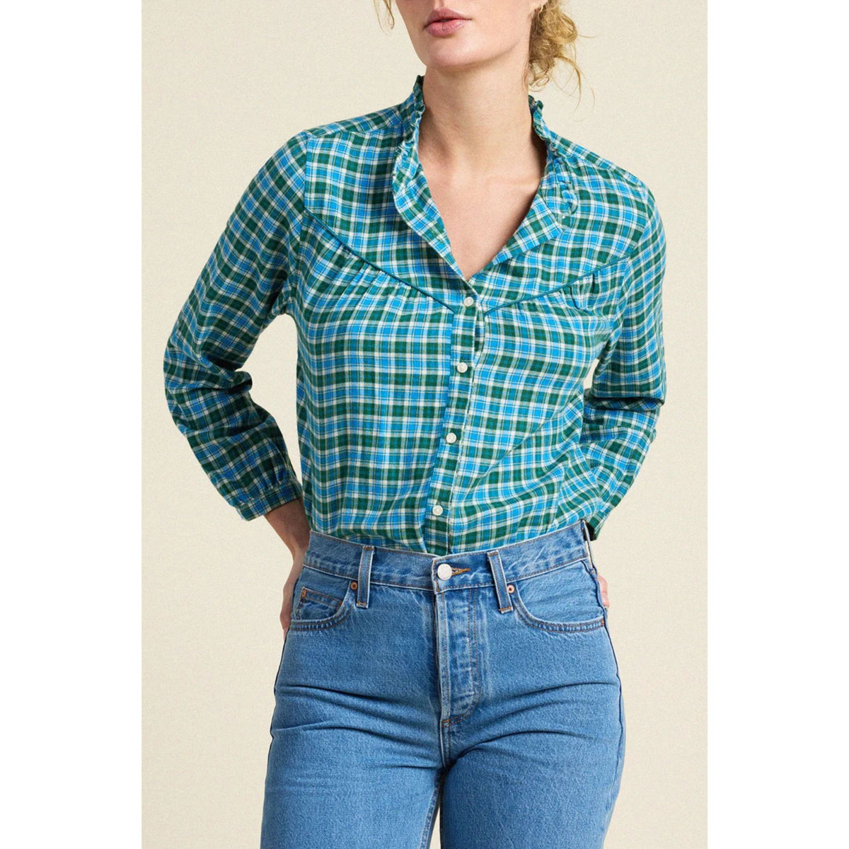 A woman wearing a green and blue plaid button-down shirt with long sleeves and blue jeans, shown from the lips down against a plain beige background.