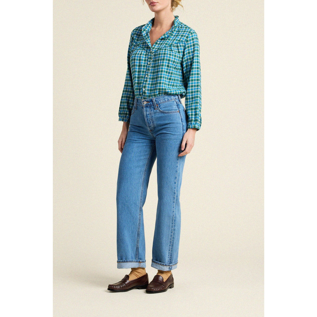 A model wearing a green and blue plaid button-down shirt tucked into light-wash blue jeans, paired with brown leather loafers, against a solid beige background.