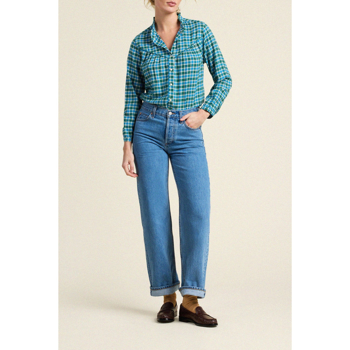 A woman modeling a green and blue plaid button-down shirt paired with light-wash blue jeans and brown loafers, standing against a solid beige studio background.