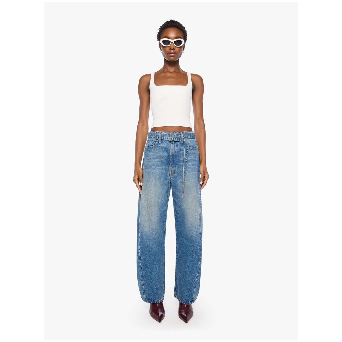 A model wearing a white ribbed crop top, wide-leg blue denim jeans, and white-framed sunglasses, standing against a light gray studio background.