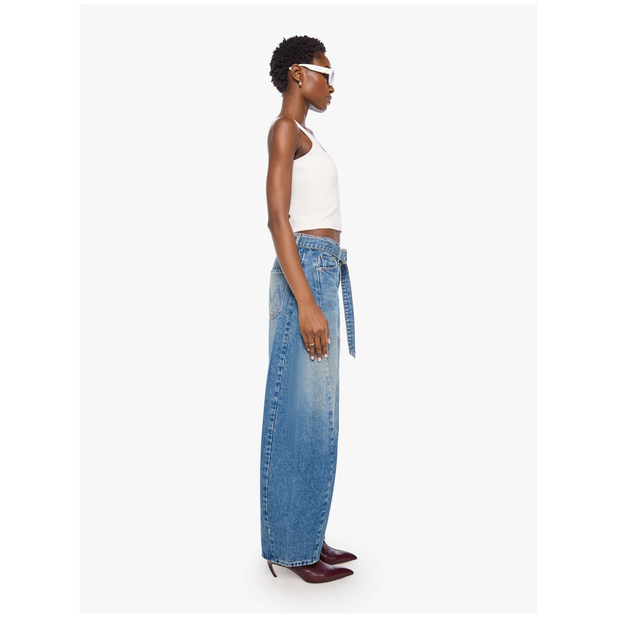 A model shown in profile wearing a white halter crop top, wide-leg blue denim jeans with a matching denim belt, and burgundy pointed-toe heels against a light gray studio background.