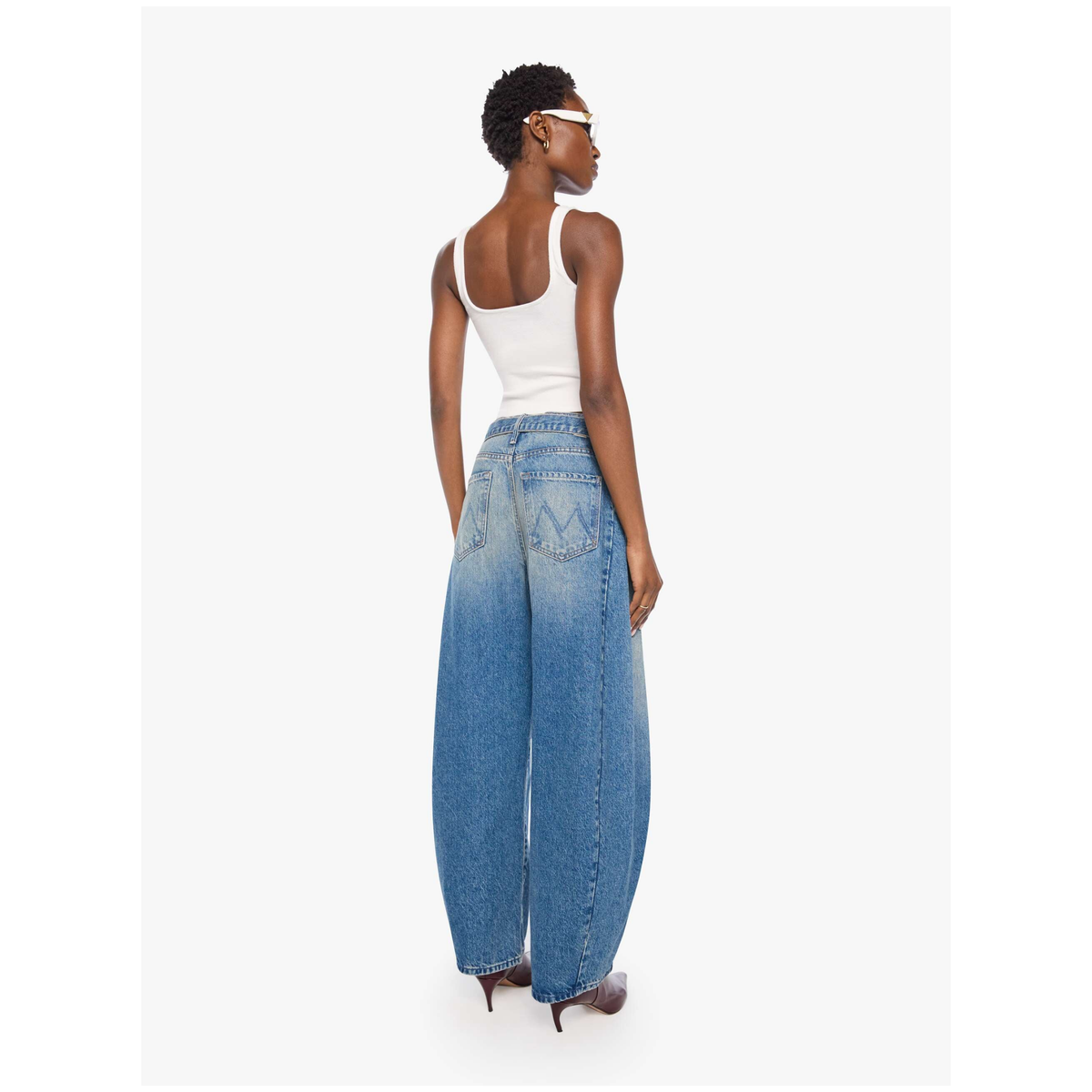 A model shown from the back wearing wide-leg blue denim jeans with a faded wash, a white tank top, and brown pointed-toe heels against a light gray studio background.