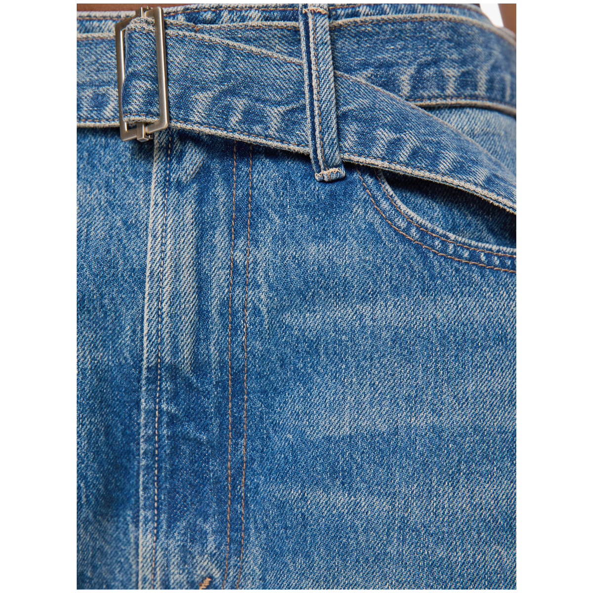 Close-up of a blue denim skirt showing the waistband with a silver-tone metal belt buckle, belt loops, front seam, and part of a pocket.