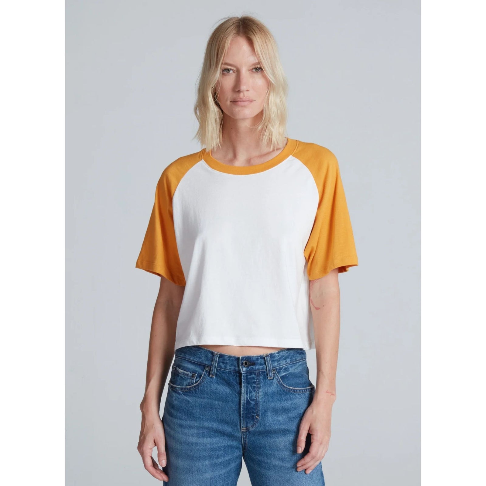 Front three-quarters view of a blonde model wearing the ASKK NY Short Sleeve Baseball Tee in Mustard—a white cropped t-shirt with mustard yellow raglan sleeves—paired with medium wash blue jeans against a light gray background.