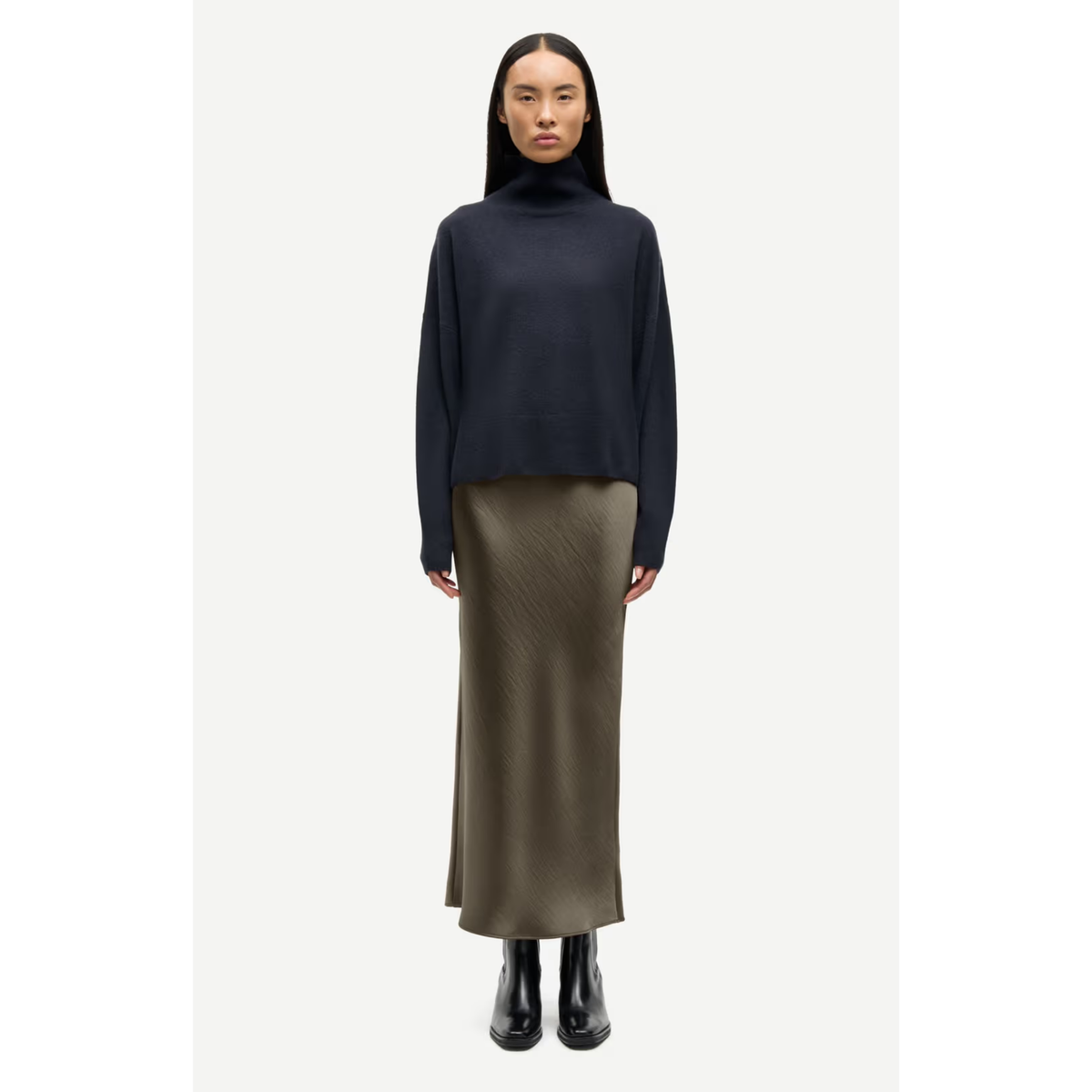 A model stands against a light gray studio background wearing a dark navy turtleneck sweater, a long olive green skirt, and black boots.