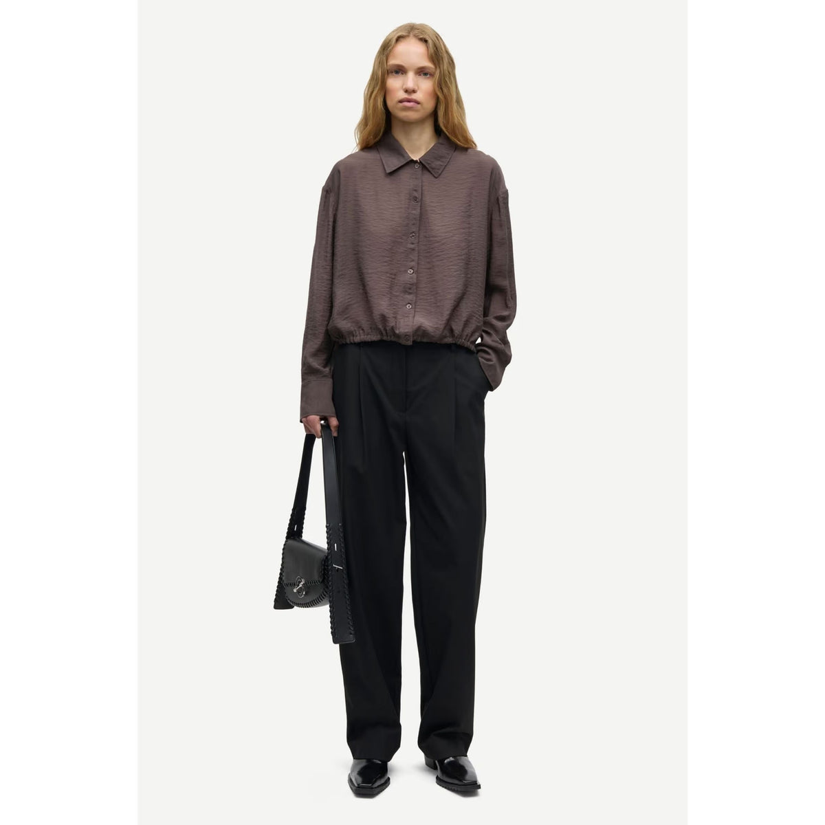 A model stands against a light gray studio background wearing a textured dark brown long-sleeve button-up shirt, black wide-leg trousers, and black shoes, while holding a small black shoulder bag.