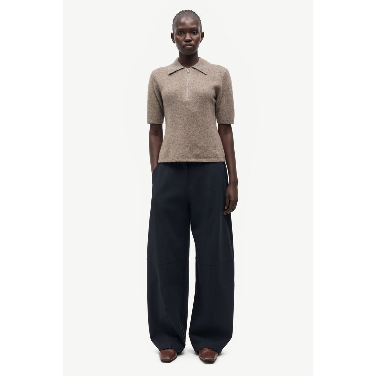 A model stands against a plain light background wearing a short-sleeve knit polo-style top in taupe and very wide-leg dark trousers.