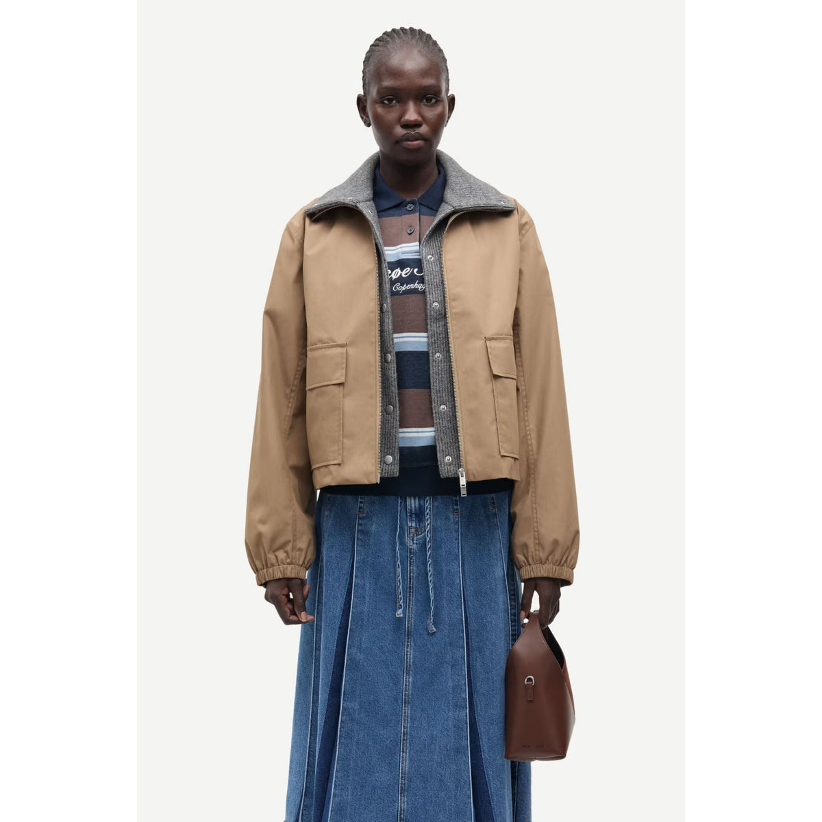 A model stands against a light gray studio background wearing a cropped tan jacket with a gray ribbed collar over a striped knit top and a long blue denim skirt, while holding a brown leather handbag.