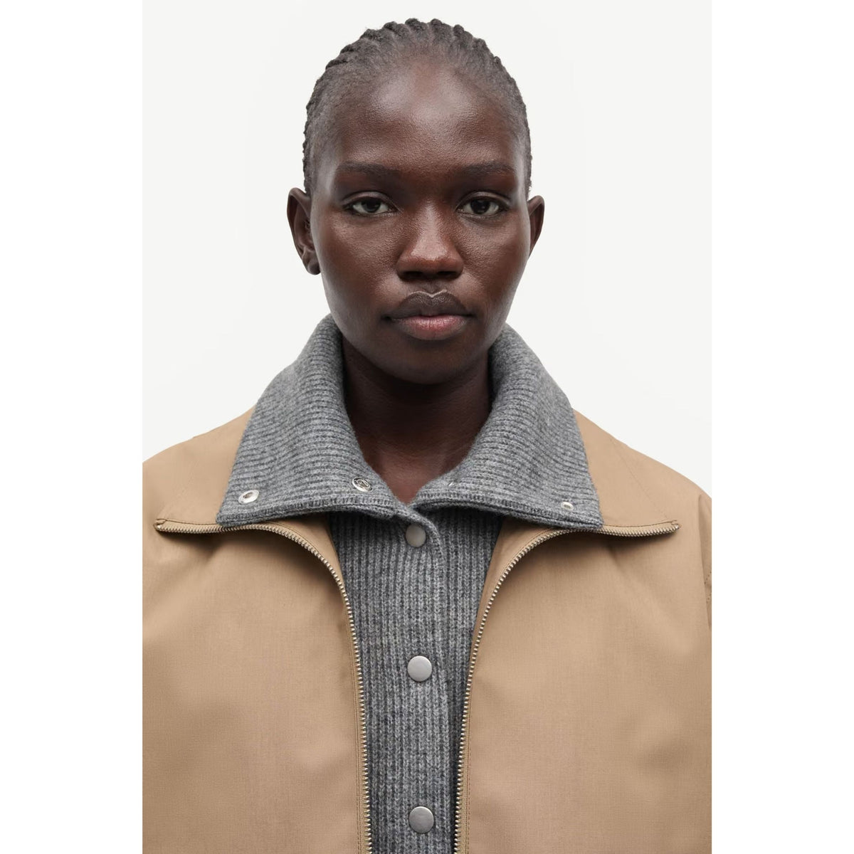 A close-up of a model wearing a tan jacket with a ribbed gray knit collar over a gray knit top, shot against a plain light background.