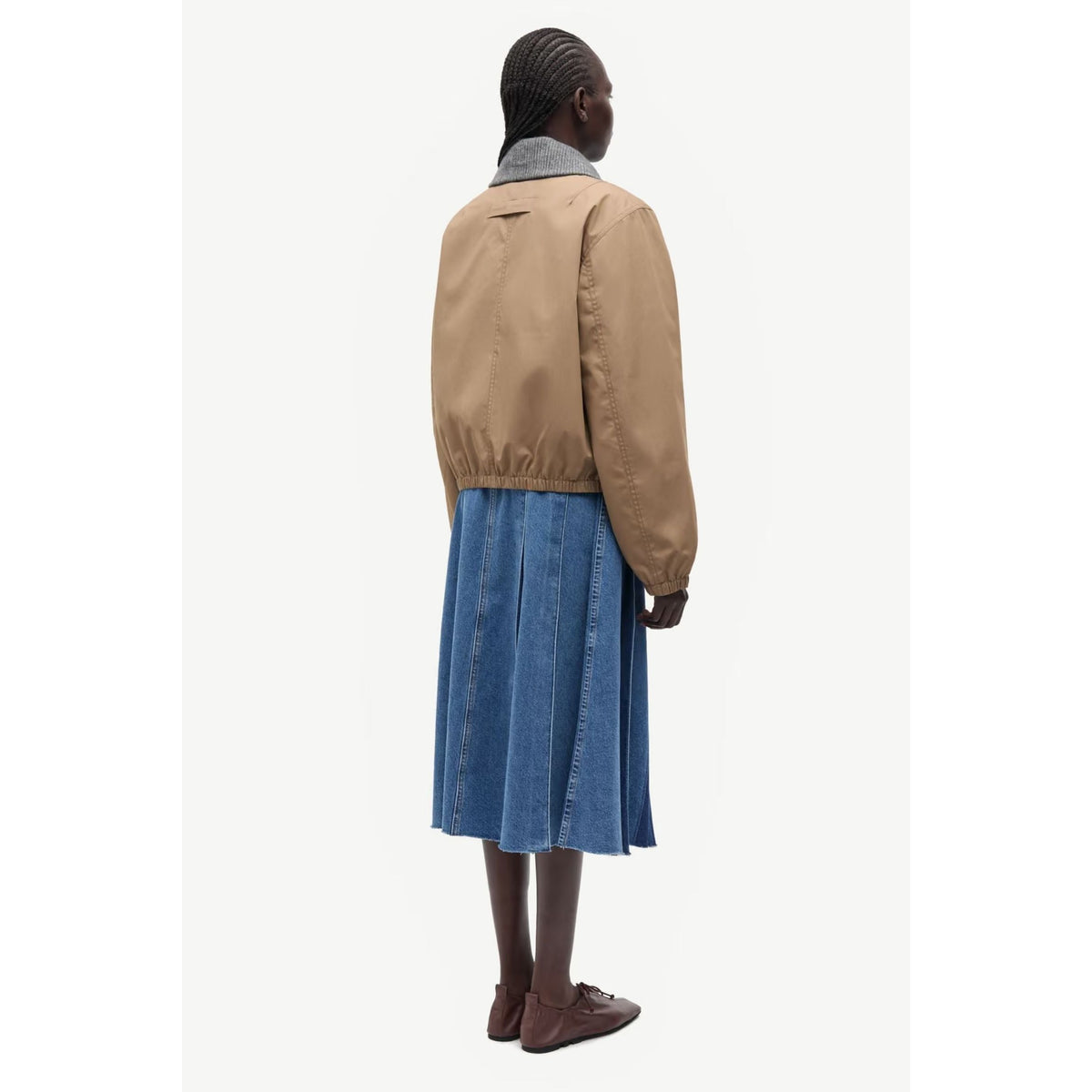 A model shown from the back wearing a tan jacket with a gray collar, a blue pleated midi skirt, and brown loafers, set against a plain off-white background.