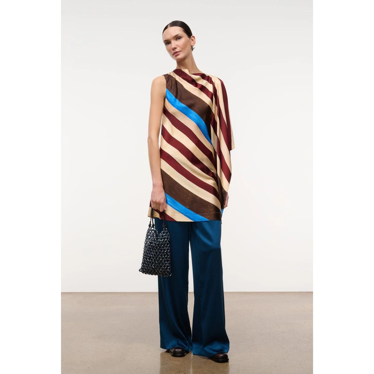 A model stands in a studio wearing a sleeveless draped top with diagonal stripes in brown, beige, burgundy, and bright blue, paired with dark teal wide-leg pants and holding a black patterned handbag.