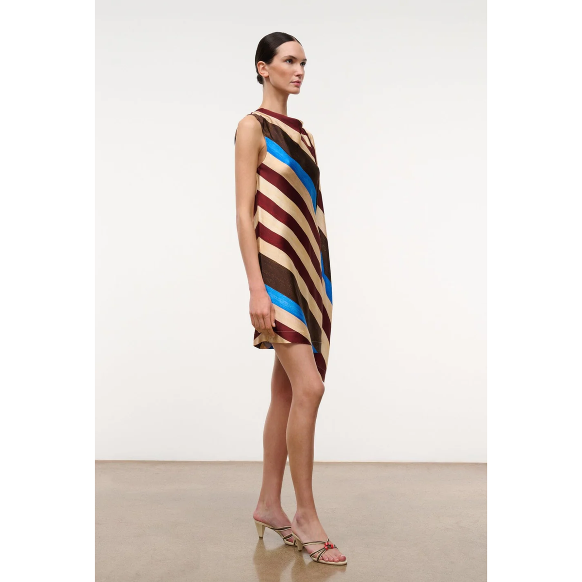 A model stands in profile wearing a sleeveless, high-neck mini dress with diagonal stripes in beige, brown, burgundy, and bright blue. She is wearing clear high-heeled mules on a minimalist studio set with a light gray wall and beige floor.