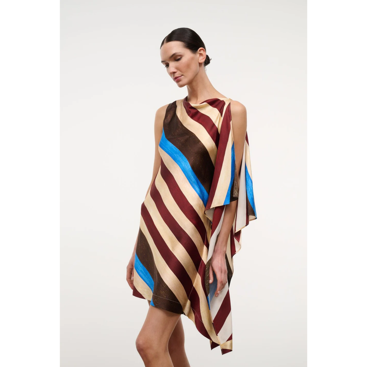 A model wearing a sleeveless, draped mini dress with diagonal stripes in beige, brown, burgundy, and bright blue, set against a plain light gray studio background.