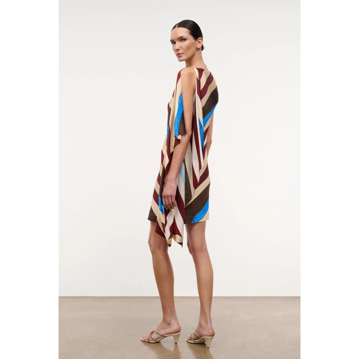 A model wearing a sleeveless, high-low dress with a bold geometric pattern in brown, beige, blue, and white, standing in profile against a neutral studio background.