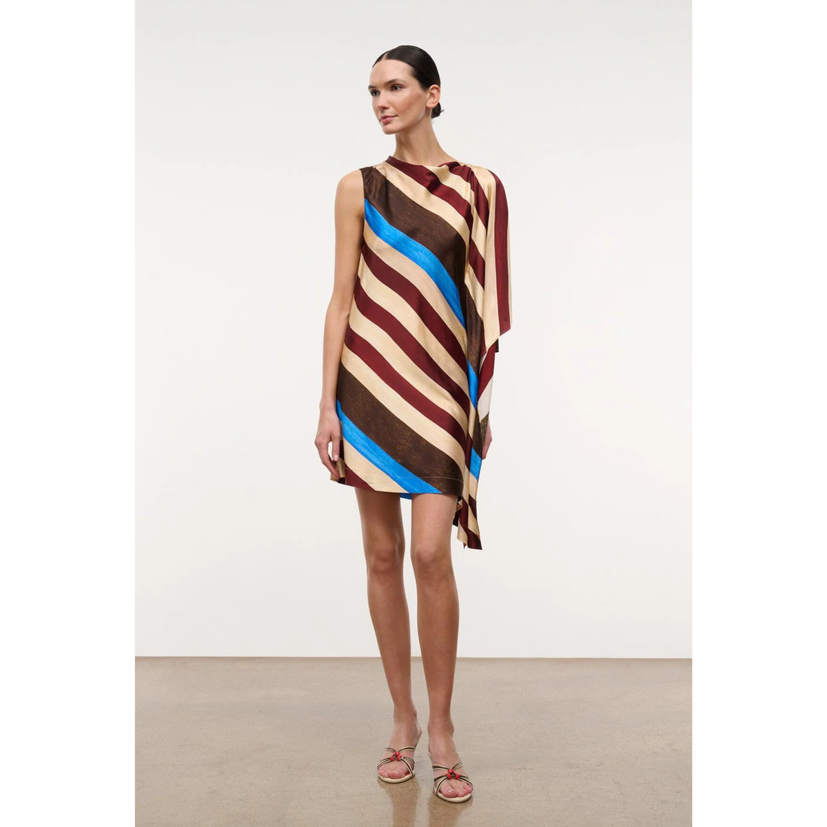 A model stands in a studio wearing a sleeveless, draped mini dress with diagonal stripes in brown, beige, burgundy, and bright blue, paired with pink sandals.