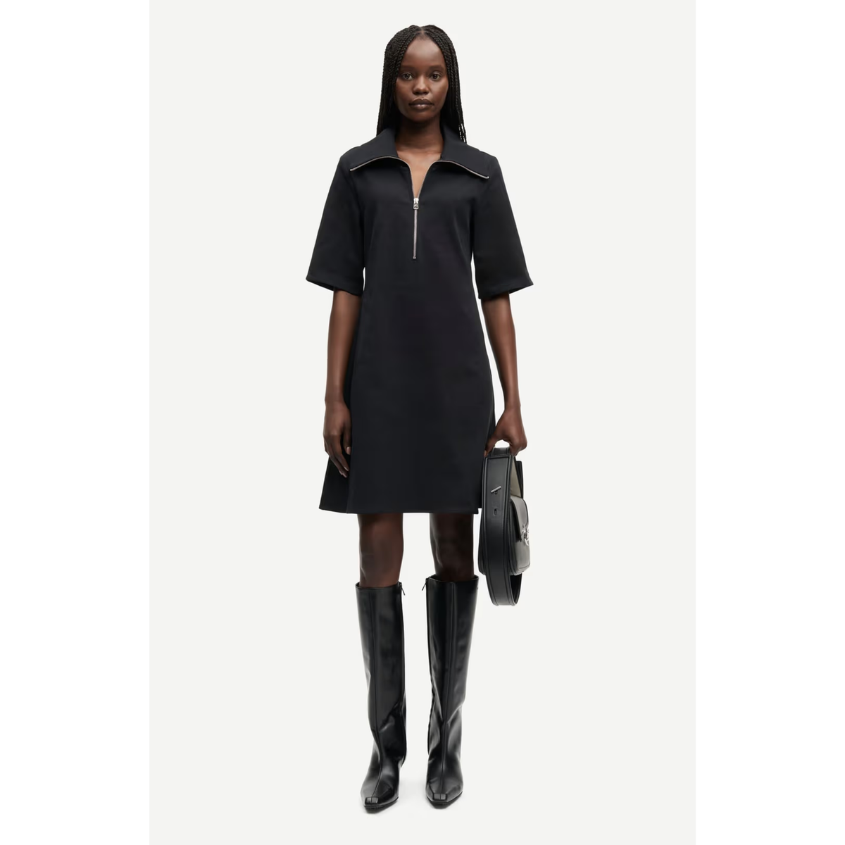 A model wearing a black short-sleeve mini dress with a half-zip neckline, paired with black knee-high boots and holding a black handbag, set against a plain light gray background.