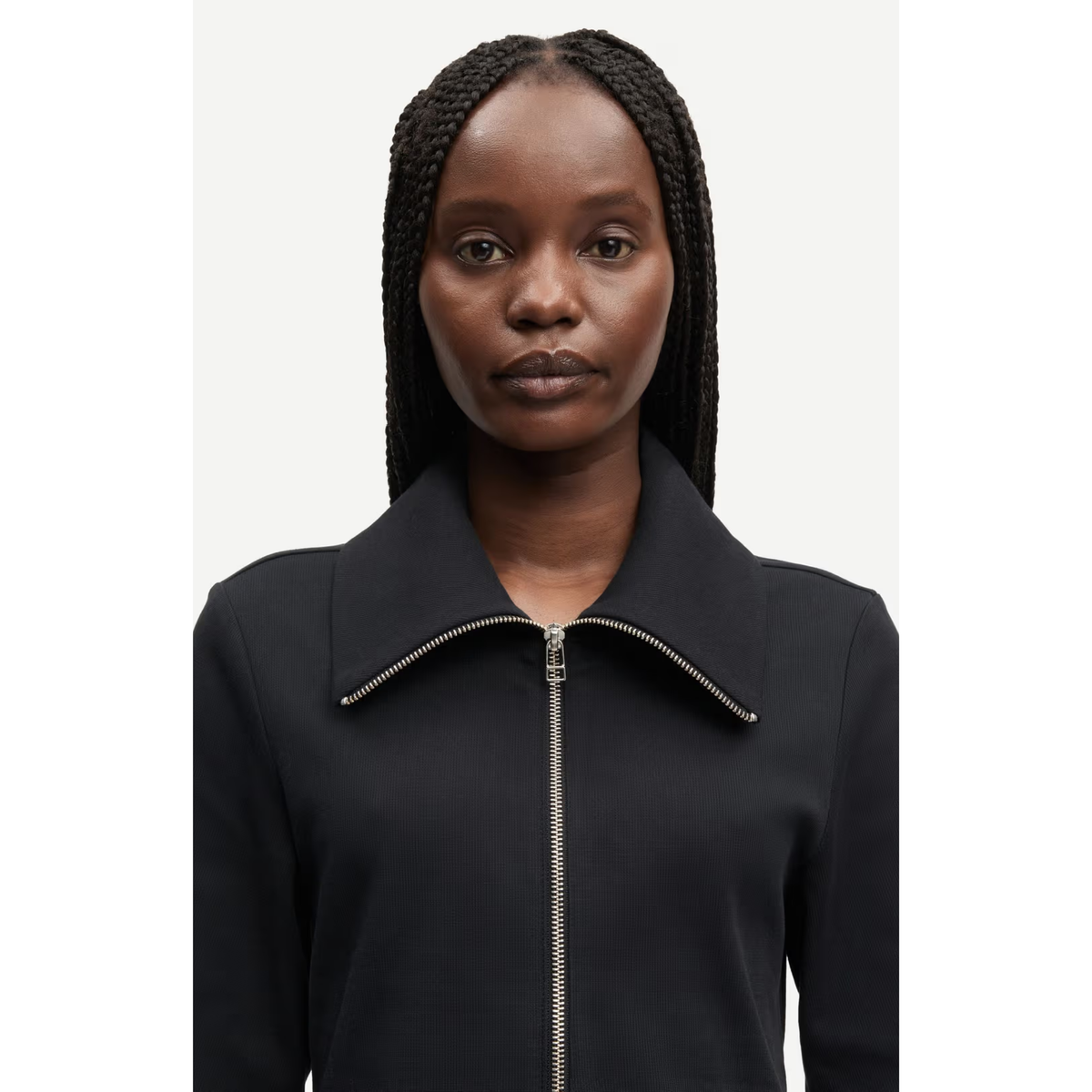 A woman with braided hair wearing a black zip-up jacket with a high collar and silver zipper trim, shown against a plain light gray background.