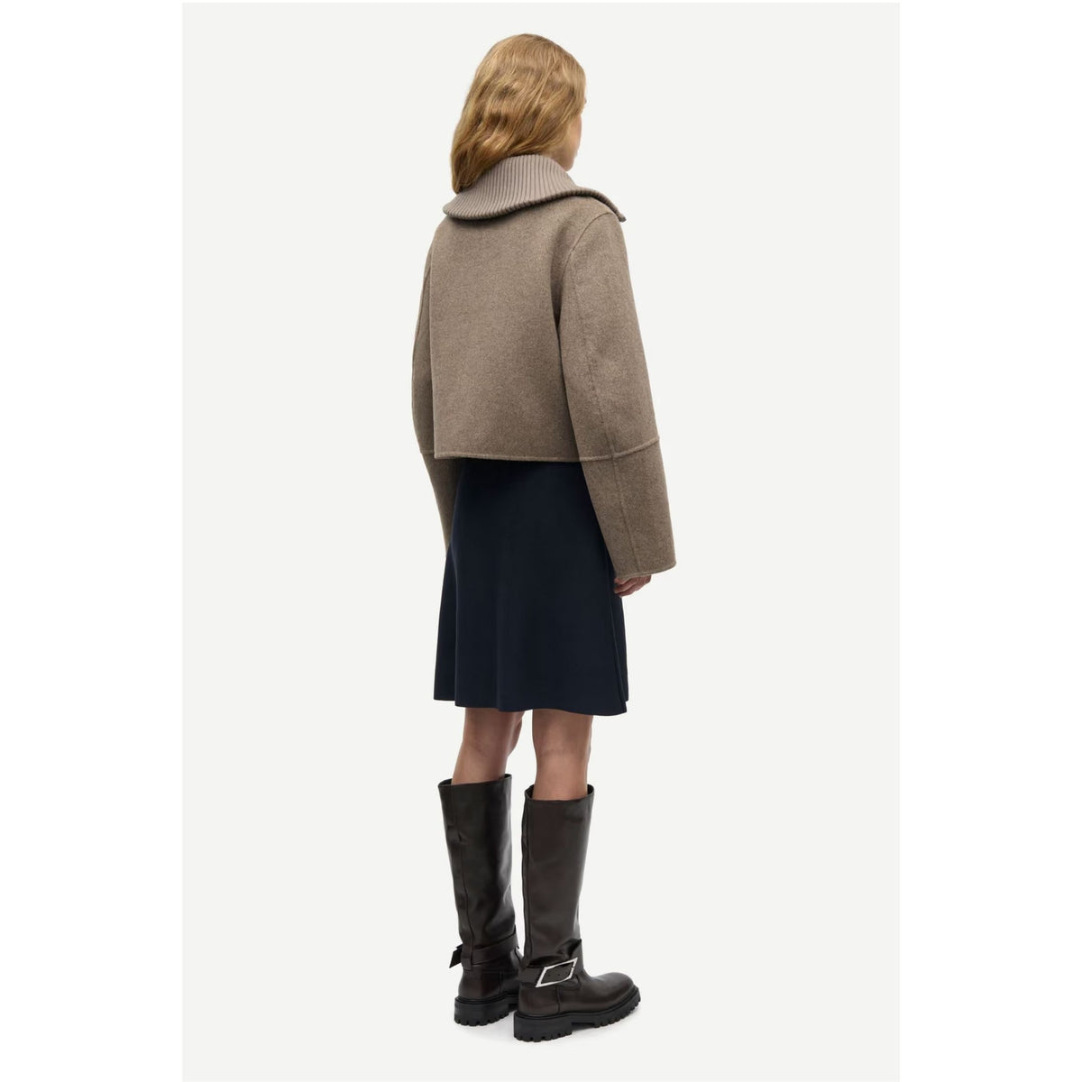 A model shown from the back wearing a cropped taupe-brown coat, a dark navy skirt, and dark brown leather boots with buckle details, set against a plain light gray background.