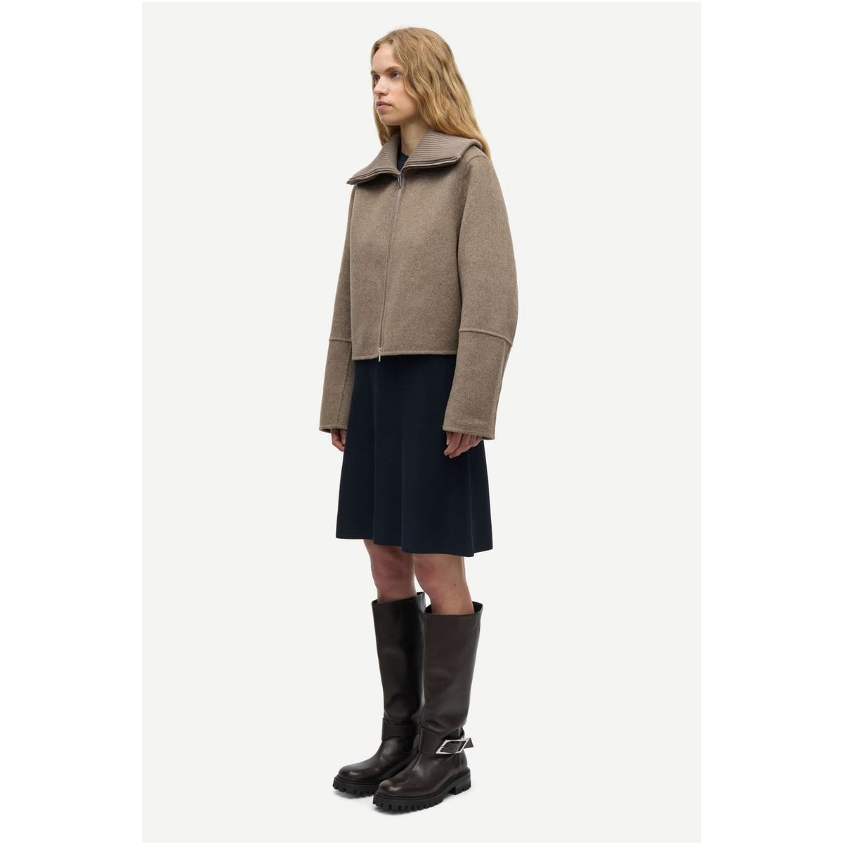 A woman stands in profile wearing a taupe cropped jacket with a wide collar, a dark navy skirt, and dark brown knee-high boots against a plain light gray studio background.