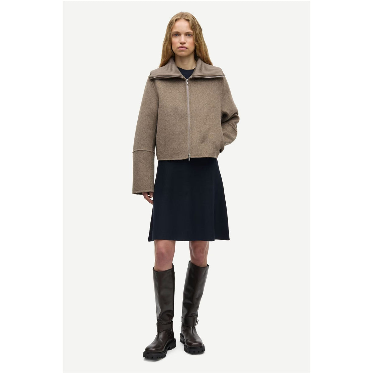 A model stands against a light gray studio background wearing a cropped taupe-brown zip-front jacket with a wide collar, a dark navy midi skirt, and tall dark brown leather boots.