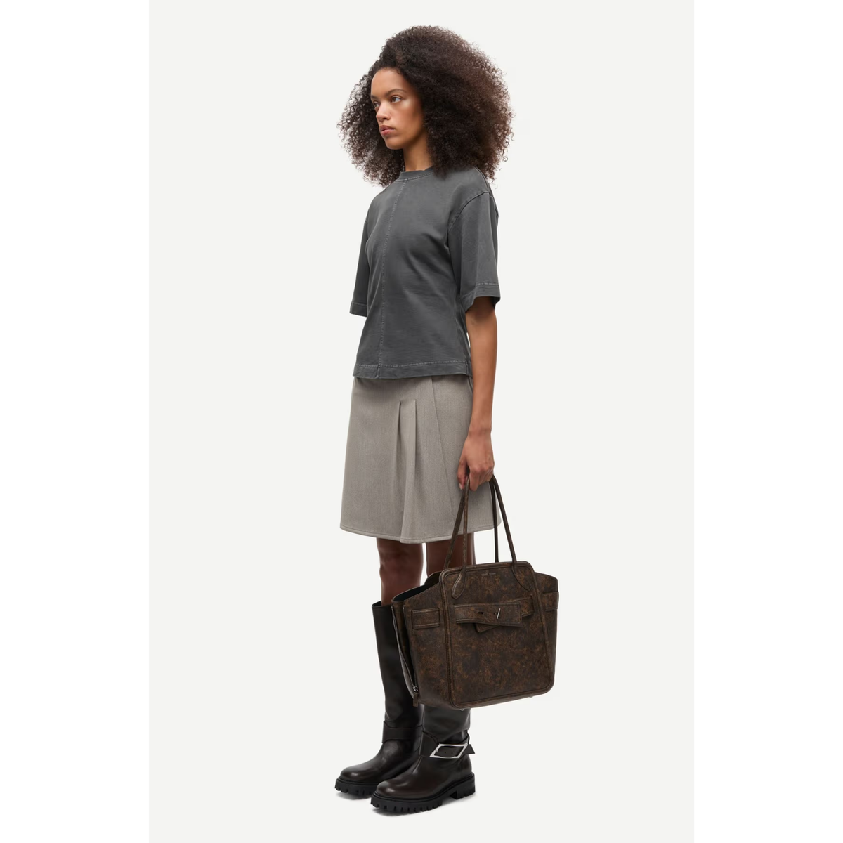 A model stands in profile wearing a dark gray cropped t-shirt, a light gray pleated skirt, and black boots, while holding a brown monogram handbag against a plain off-white background.