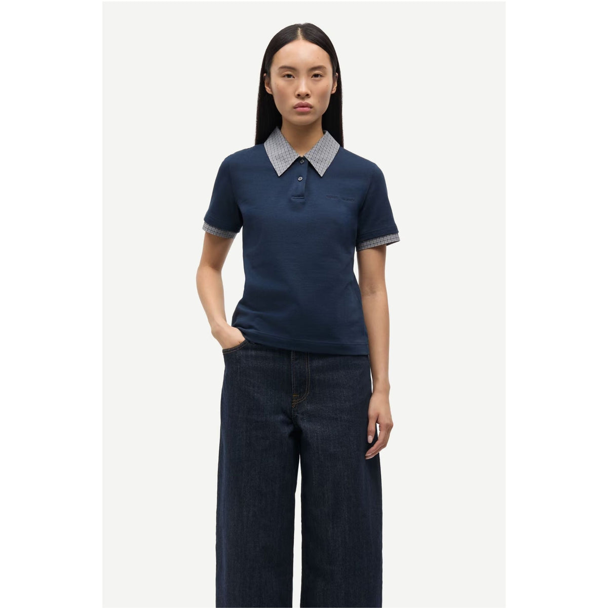 A woman modeling a navy blue short-sleeve polo shirt with a contrasting light gray and white patterned collar and sleeve cuffs, paired with dark wide-leg jeans against a light gray studio background.