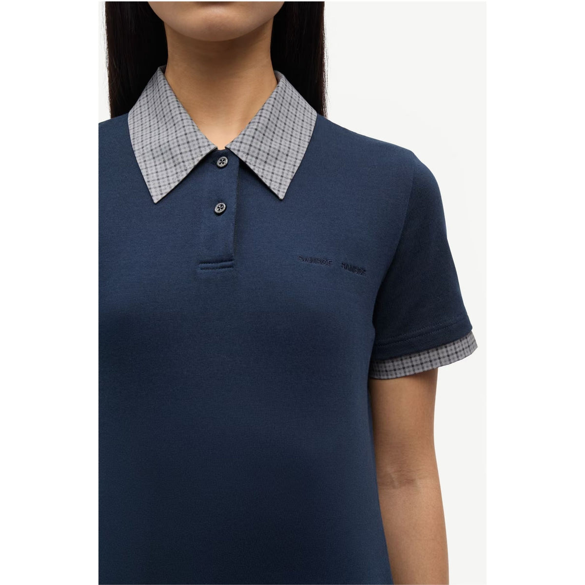 A close-up of a woman wearing a navy blue polo shirt with a light gray and white checkered collar and matching checkered sleeve cuffs. The brand name 'SAMSOE SAMSOE' is subtly embroidered on the chest.