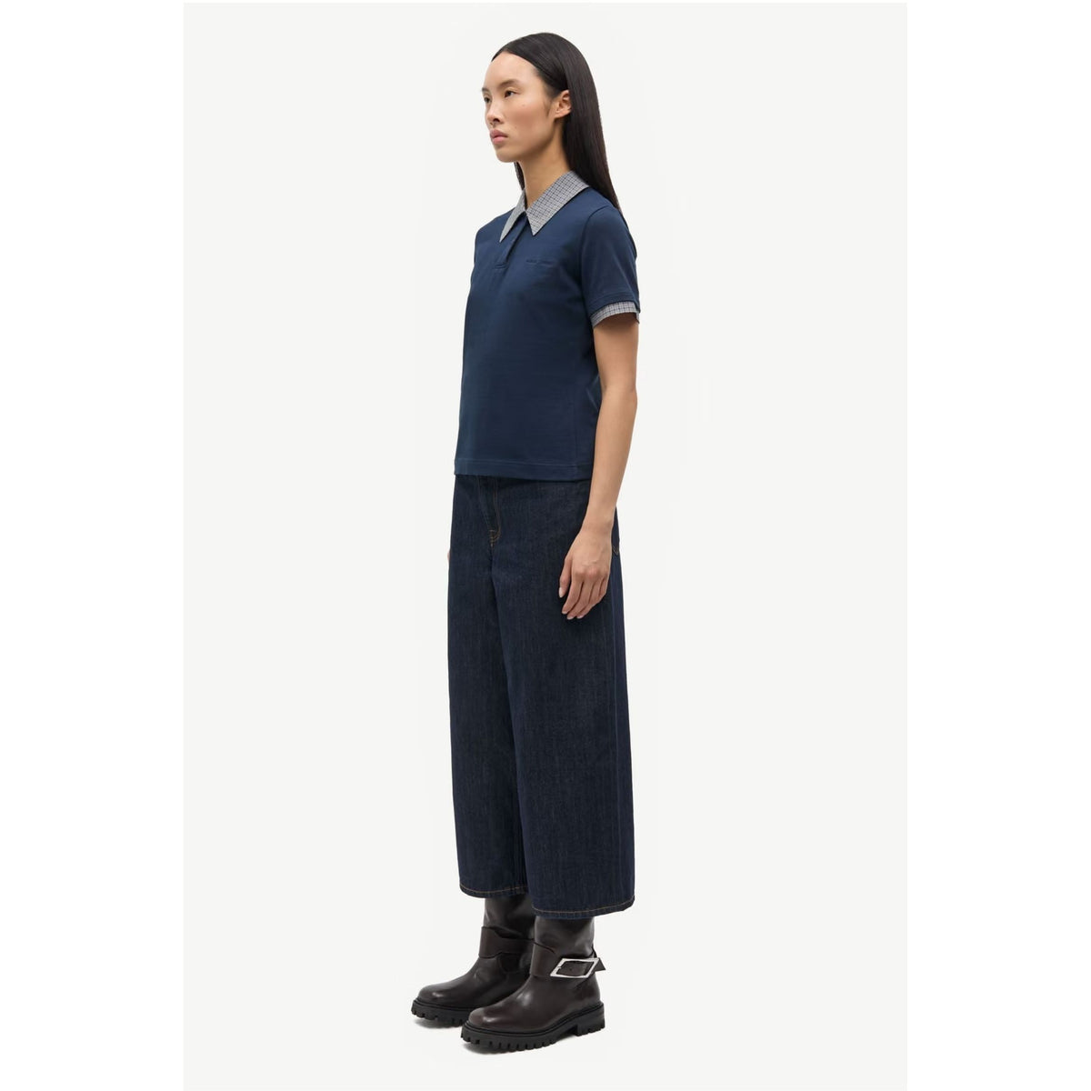 A woman stands in profile wearing a dark navy short-sleeve polo shirt with a contrasting light gray collar and cuffs, paired with dark wide-leg pants and black ankle boots, against a plain light gray studio background.