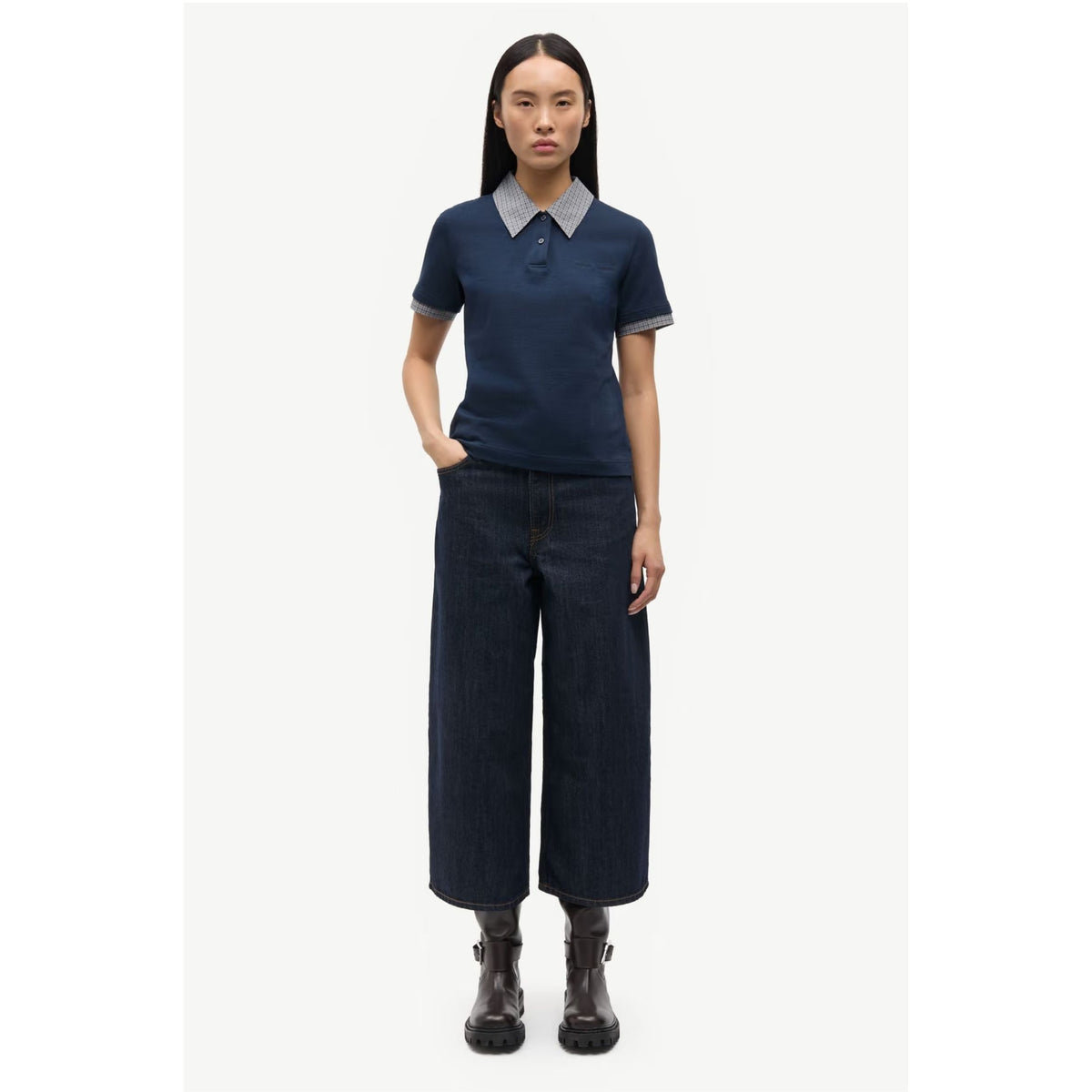 A model stands against a light gray studio background wearing a navy short-sleeve polo shirt with a contrasting light gray collar and cuffs, paired with dark wide-leg cropped pants and black ankle boots.