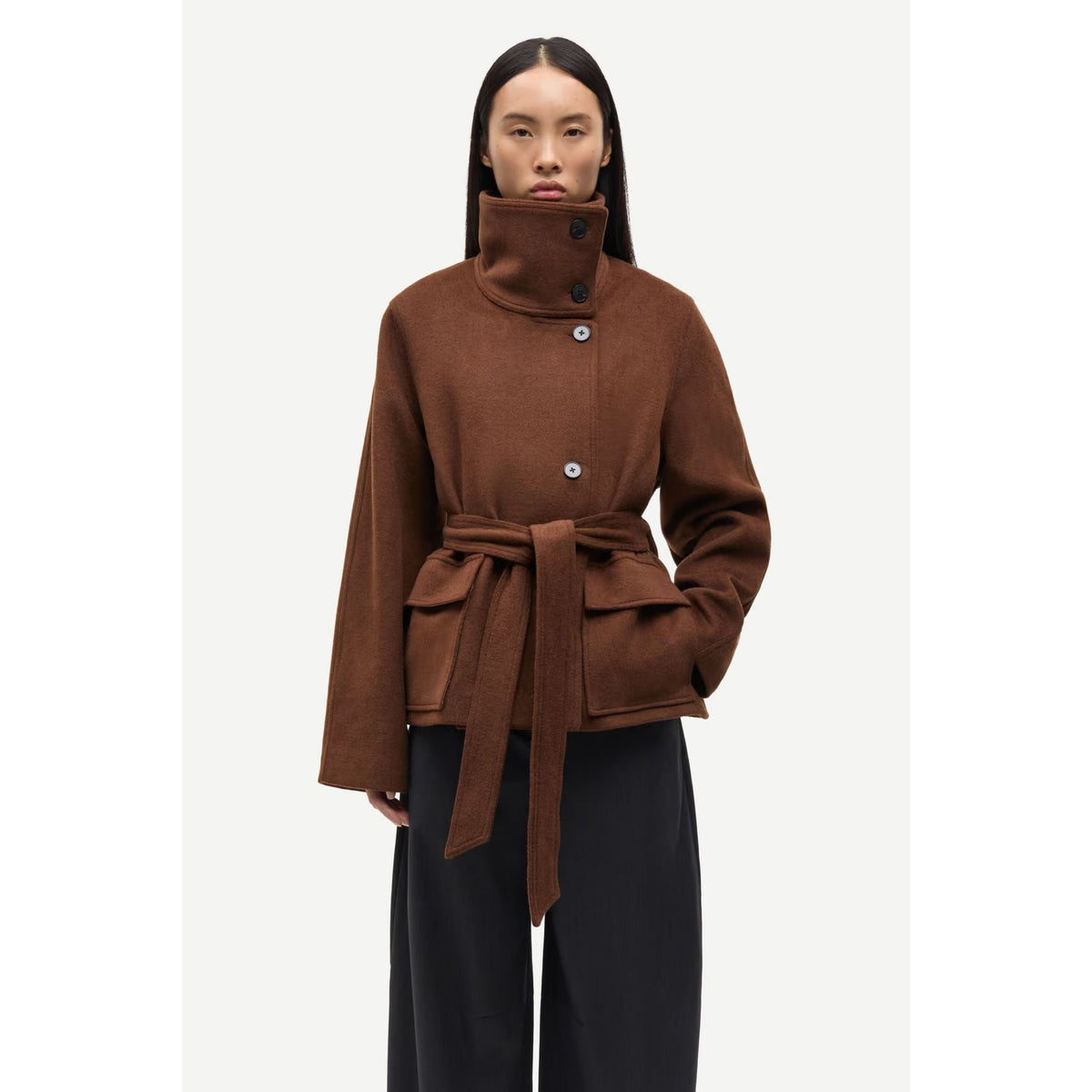 A model wearing a brown high-neck belted coat with front buttons and a tie belt, paired with black wide-leg trousers, against a light gray studio background.