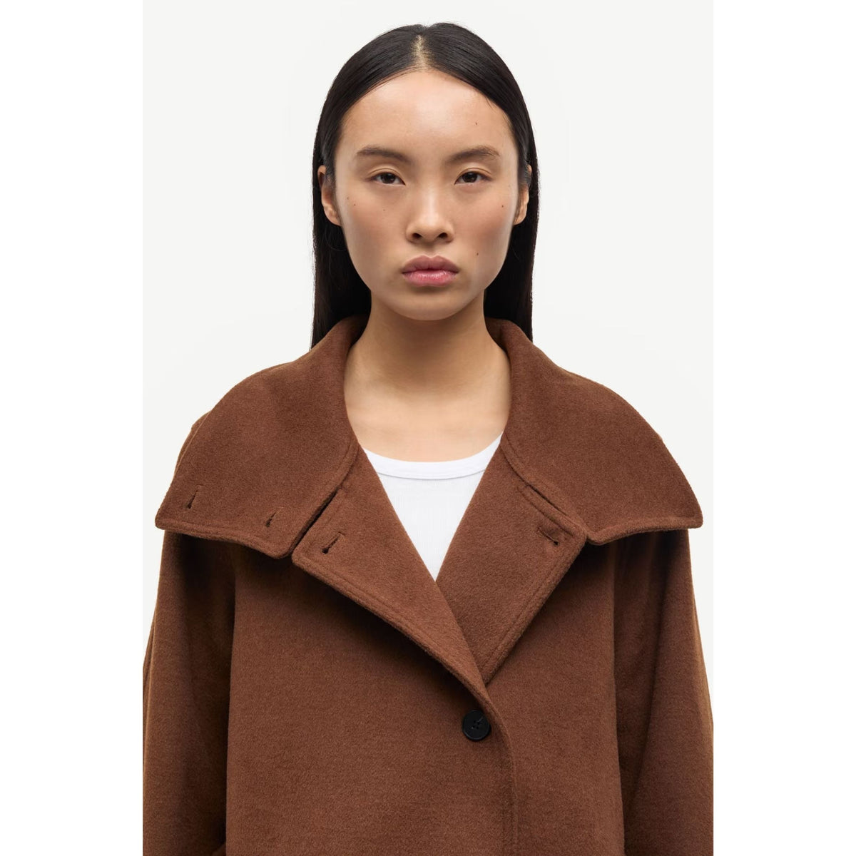 A model wearing a brown wool-blend coat with a wide collar and a single dark button, layered over a white top, shot against a plain light gray background.