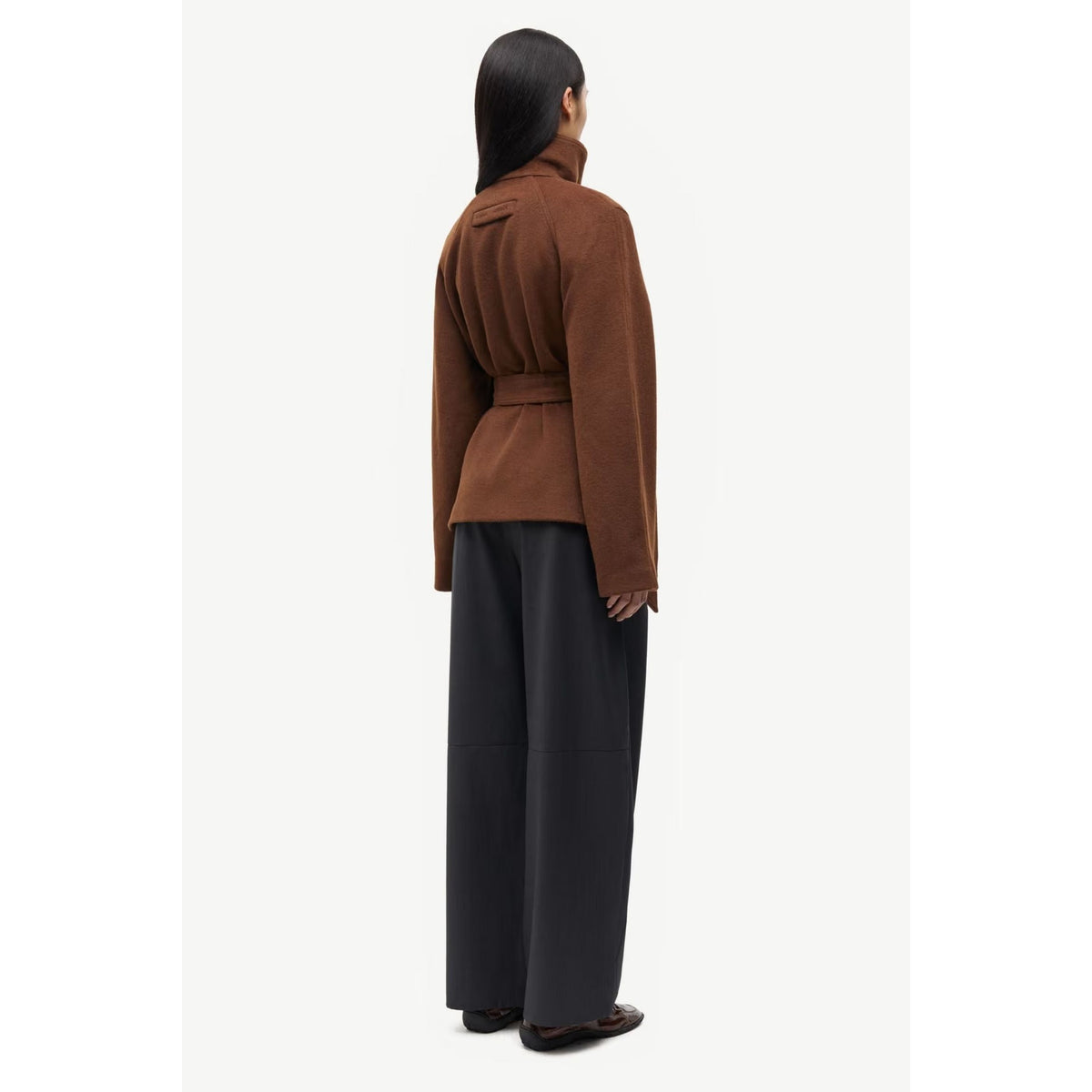 A model shown from the back wearing a brown belted wrap jacket and dark charcoal wide-leg trousers, standing against a plain off-white background.