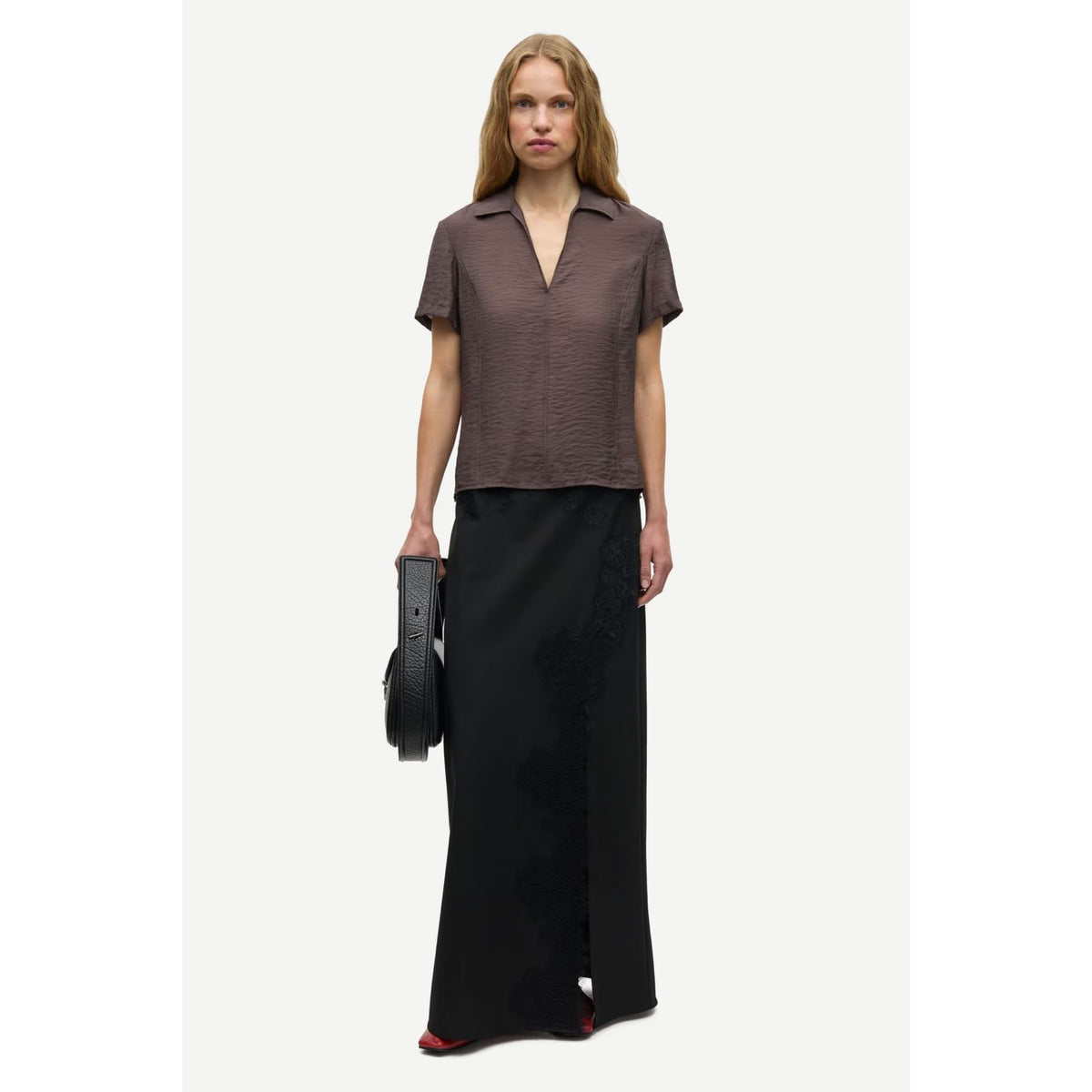 A model stands against a light gray studio background wearing a short-sleeve textured brown collared top and a long black skirt, holding a black handbag.