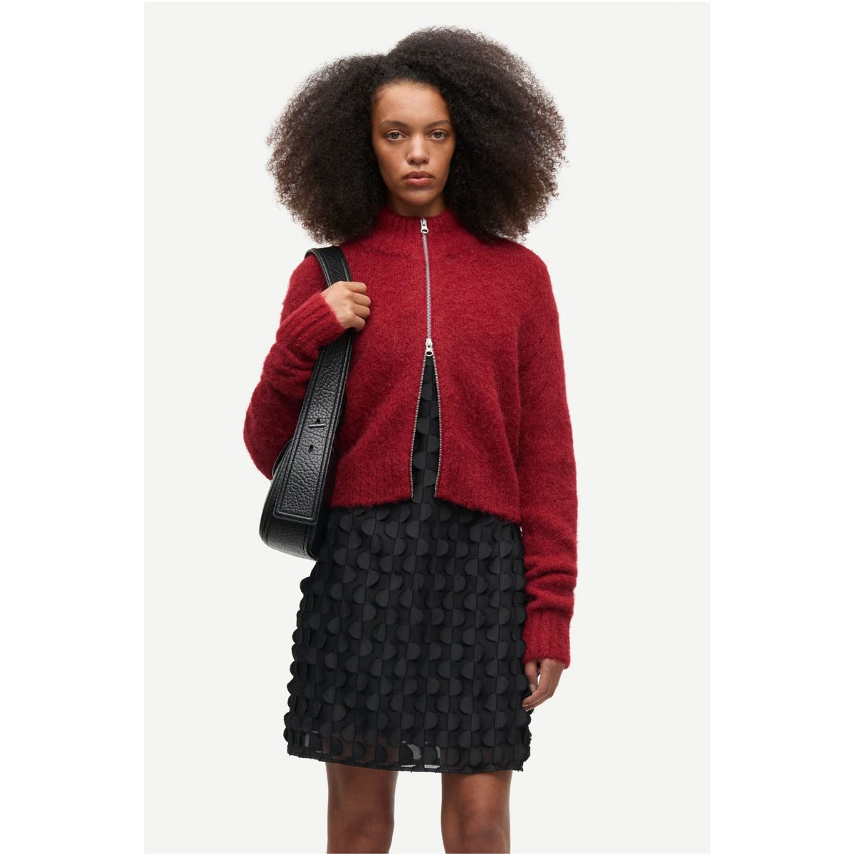 A model with dark curly hair is wearing a red fuzzy zip-up mock neck sweater and a black textured skirt, carrying a black shoulder bag against a light gray background.