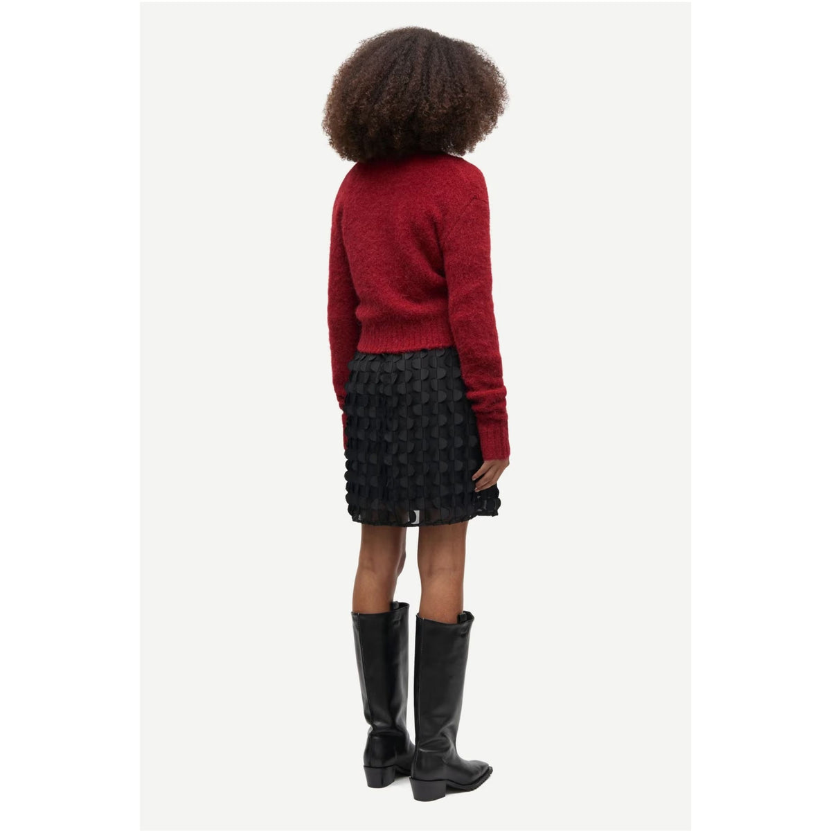 A child model shown from the back wearing a red knit sweater, a black textured mini skirt, and black knee-high boots against a light gray studio background.