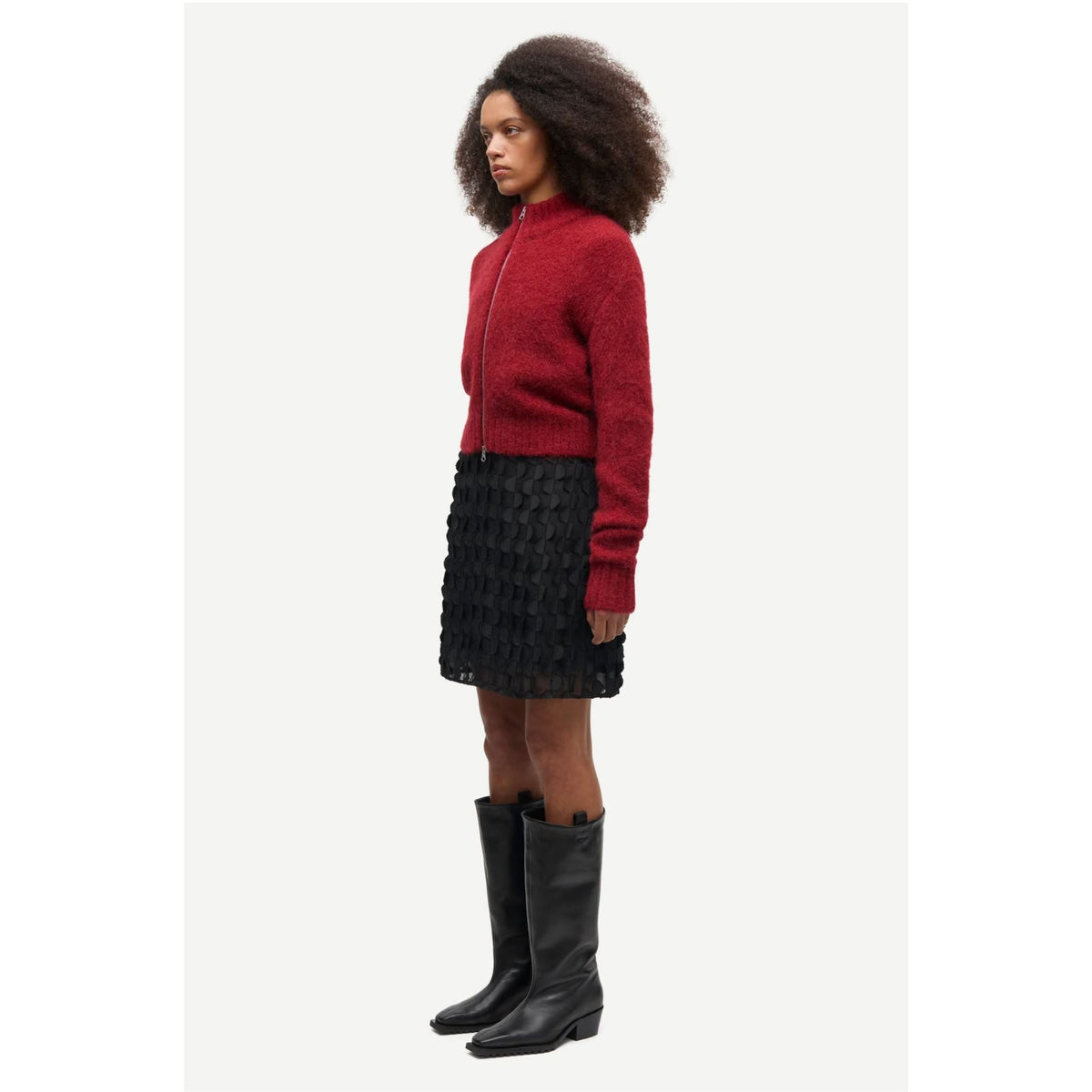A model is shown in profile wearing a red knit zip-up sweater, a black textured mini skirt, and black leather knee-high boots against a light gray background.