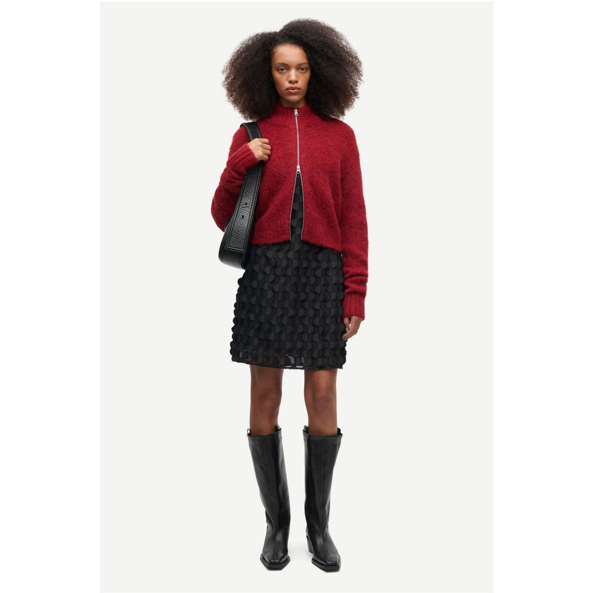 A model with a large dark afro is wearing a red cropped zip-up sweater, a black textured mini skirt, and black knee-high boots, while carrying a black shoulder bag against a light gray studio background.