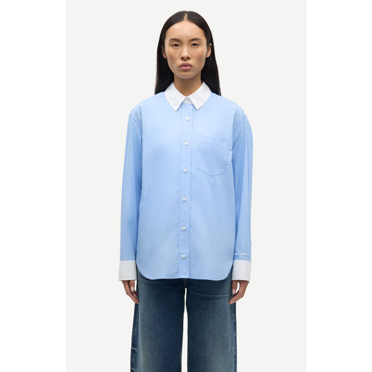 A model wearing a light blue long-sleeve button-up shirt with a contrasting white collar and cuffs, paired with dark blue jeans, set against a plain light gray background.