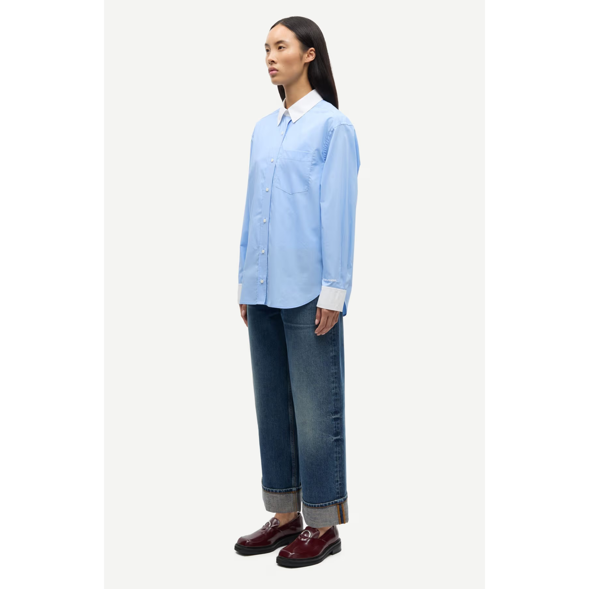 A model is shown in profile wearing a light blue long-sleeve button-up shirt with a white collar, dark blue wide-leg jeans, and dark red leather shoes, set against a plain off-white studio background.