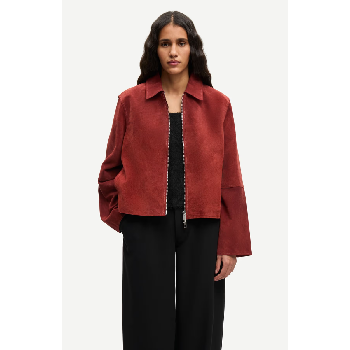A woman modeling a cropped, long-sleeve collared jacket in a rust red suede-like material with a front zipper, worn over a black top and black wide-leg pants against a light gray studio background.