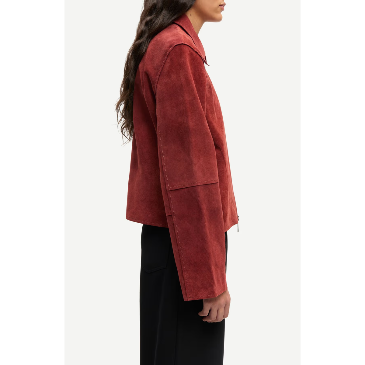 A side-profile view of a model wearing a rust-red suede jacket and black wide-leg trousers against a clean white background.