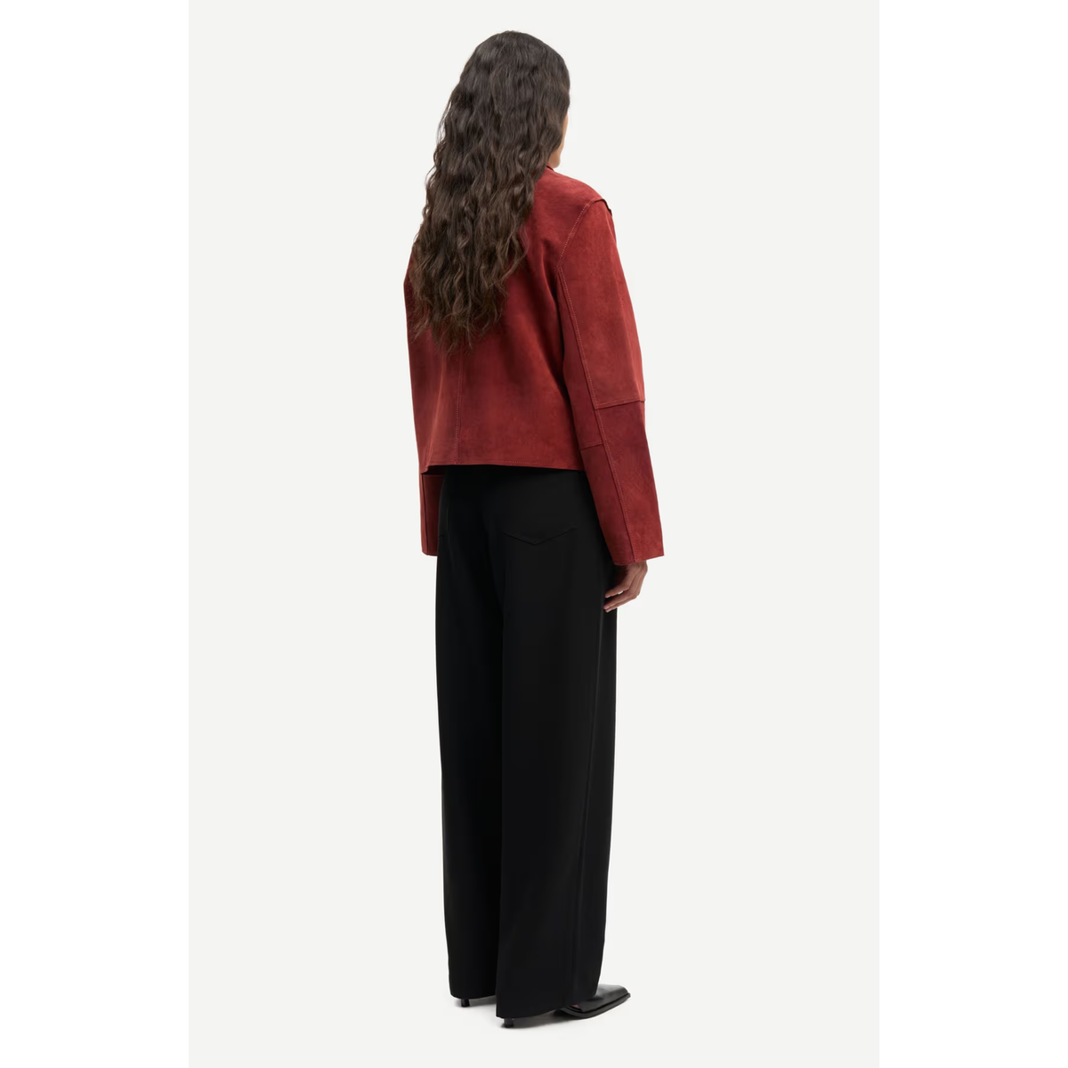 A model shown from the back wearing a rust-red cropped jacket with wide sleeves and black wide-leg trousers, set against a plain off-white studio background.