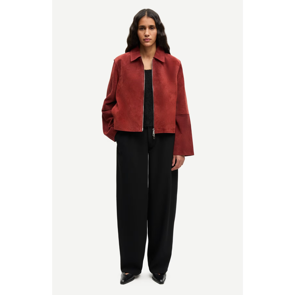A model stands against a light gray studio background wearing a cropped, long-sleeve rust-red suede jacket over a black top and black wide-leg trousers.